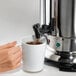 A hand pouring coffee from a Hamilton Beach stainless steel coffee urn into a mug.