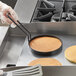 A hand in a glove holding a Vigor egg ring over a round black pan with a pancake in it.