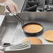 A hand in a glove using a Vigor egg ring to cook a pancake in a pan.
