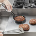 A hand using a black metal Vigor egg ring to cook hamburger patties on a pan.