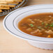 A Fineline white plastic soup bowl with silver bands filled with soup and bread sticks.