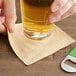 A hand holding an EcoChoice natural kraft beverage napkin with a glass of beer on a table in a bar.