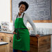 A smiling woman wearing a Choice Kelly Green poly-cotton bib apron with black webbing accents standing in front of a counter.