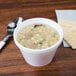 A white foam bowl of soup with a spoon and crackers on a table.