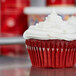 A red cupcake with cream cheese frosting in a Hoffmaster white fluted baking cup.