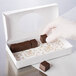 A gloved hand holding a white candy box with a clear oval window filled with chocolate pieces.