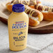 A close-up of a Grey Poupon Dijon Mustard squeeze bottle next to hot dogs.