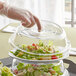 A hand wearing plastic gloves lifts a clear polycarbonate plate cover off a salad.
