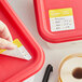 A hand labeling a red container with a Noble Products Tuesday 2" x 2" Removable Day of the Week label.