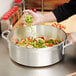 A person wearing gloves and an apron putting baby carrots into a Vollrath Wear-Ever aluminum brazier pan on a counter.