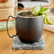 An Acopa Alchemy black Moscow mule mug filled with ice and lime slices on a marble coaster.