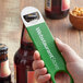 A hand using a green Webstaurant bottle opener to open a brown bottle.