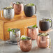 An Acopa Alchemy copper Moscow Mule mug filled with ice and a lime leaf on a table with a group of mugs.