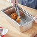 A person using tongs to serve fried chicken in a stainless steel pan.