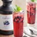 A glass of blueberry lemonade with mint leaves next to a bottle of Capora Blueberry Flavoring Sauce.