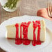 A white plate with a slice of cheesecake topped with red Capora strawberry flavoring sauce.