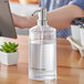 A clear plastic round hand sanitizer dispenser with a stainless steel pump on a table.