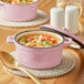 A Valor Himalayan Salt enameled cast iron pot with pink soup inside and a lid on a table with a bowl of soup and a spoon.