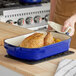 A person holding a blue Valor enameled cast iron roasting pan with chicken on a table in a home kitchen.