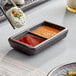 A black rectangular Acopa stoneware dish with 2 compartments holding sushi and sauce.