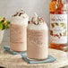 A bottle of Monin Premium Hazelnut Flavoring Syrup next to two glasses of chocolate milkshakes with whipped cream and syrup.