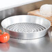 An American Metalcraft silver metal pizza pan with holes in it next to a ball of dough and tomatoes.