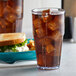 A clear plastic tumbler filled with ice and soda on a table with a sandwich.