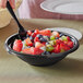 A Visions black plastic bowl filled with fruit salad with a spoon in it.