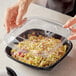 A hand reaching for a clear plastic dome lid on a Visions square bowl full of food.
