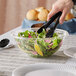 A hand using black tongs to serve salad from a Visions clear plastic bowl.