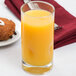 A Libbey customizable beverage glass filled with orange juice on a table with a donut and a blurry fork.