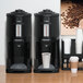 A Zojirushi stainless steel thermal gravity beverage dispenser on a table with a coffee machine and cups.