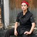 A woman wearing an Uncommon Chef black short sleeve chef coat sitting on a wooden bench.