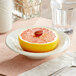 An Acopa ivory stoneware bowl with a grapefruit half in it on a table with a spoon and a glass.