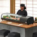 A Emperor's Select countertop refrigerated sushi display case on a counter in a sushi bar with a woman standing behind it.