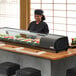A woman standing behind a countertop Emperor's Select refrigerated sushi display case.