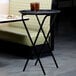 A black Lancaster Table & Seating folding tray stand with two glasses of liquid on it.