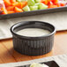 A glossy black fluted Acopa stoneware ramekin filled with white sauce on a table.