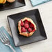 A black Acopa stoneware plate with raspberry tartlets and raspberries.