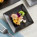A matte black Acopa square stoneware plate with food and a fork on a table.