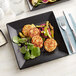 An Acopa Matte Black stoneware plate with two fish cakes, a fork, and a knife on a blue tablecloth.