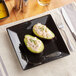 A black square stoneware plate with avocado halves and a fork on it.