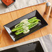A glossy black rectangular stoneware platter with a plate of asparagus and sauce and a glass of beer on a table.