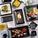 A variety of Acopa matte black square stoneware plates with food on them on a table.