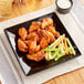 A glossy black square Acopa stoneware plate with chicken wings, celery, and carrots.