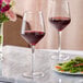 Two Acopa Silhouette wine glasses filled with red wine on a table next to a plate of food.