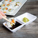 A hand holding chopsticks over a sushi roll on a white dish with dipping sauce.