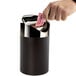 A hand putting a packet into a black and silver Cal-Mil round counter trash bin.