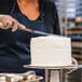 A woman in a black apron using a knife to cut a cake with Rich's Bettercreme Vanilla Whipped Icing on it.