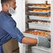 A man in a blue apron is opening a clear door on a ServIt holding cabinet to put food on a tray.